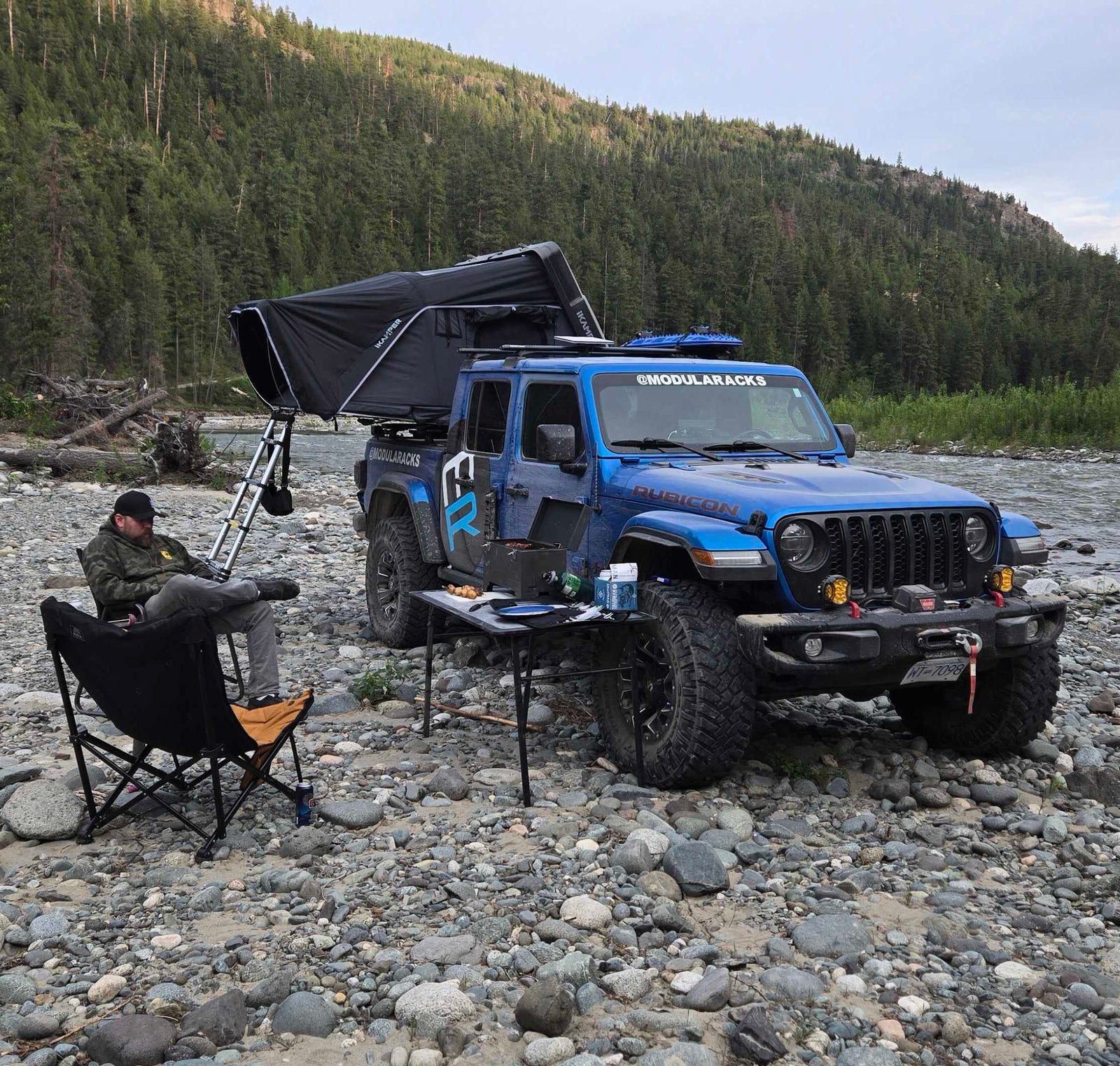 Blue Jeep Gladiator parked on a rocky riverbank with an iKamper Skycamp 4.0 Duo rooftop tent set up. A man relaxes in a camp chair next to a cooking table, with a river and forest in the background.